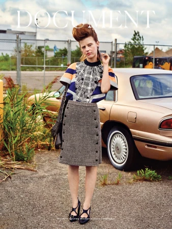 Editorial fashion portrait of a young woman wearing a skirt and top, standing near a vintage car with an urban industrial background.