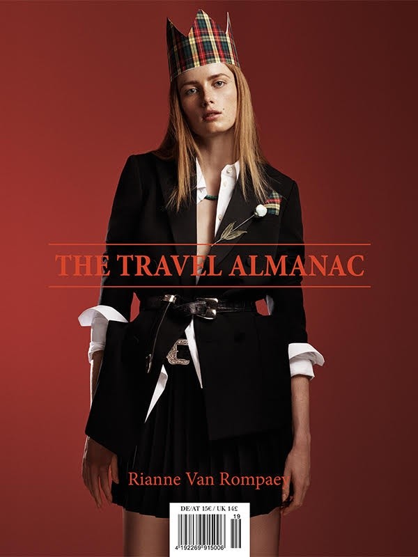Fashion editorial featuring a woman wearing a black blazer and kilt-style skirt, adorned with a tartan crown, posing against a red backdrop.