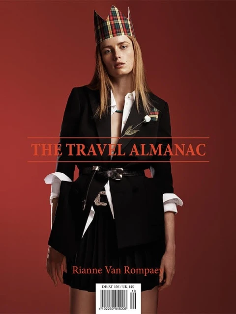 Fashion editorial featuring a woman wearing a black blazer and kilt-style skirt, adorned with a tartan crown, posing against a red backdrop.