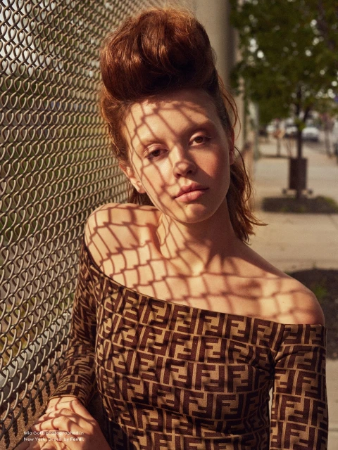 Woman poses in designer patterned off-the-shoulder top in front of chain-link fence with tree-lined street in soft outdoor light.