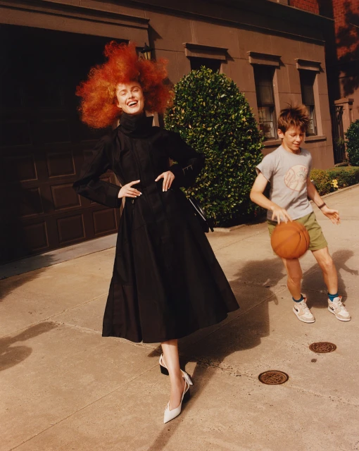 A young redhead woman in a black dress poses on a city street while a boy dribbles a basketball in a lifestyle shot.