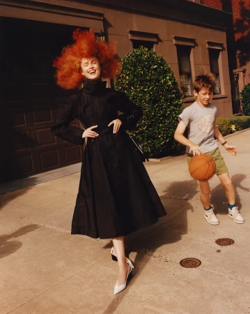A young redhead woman in a black dress poses on a city street while a boy dribbles a basketball in a lifestyle shot.