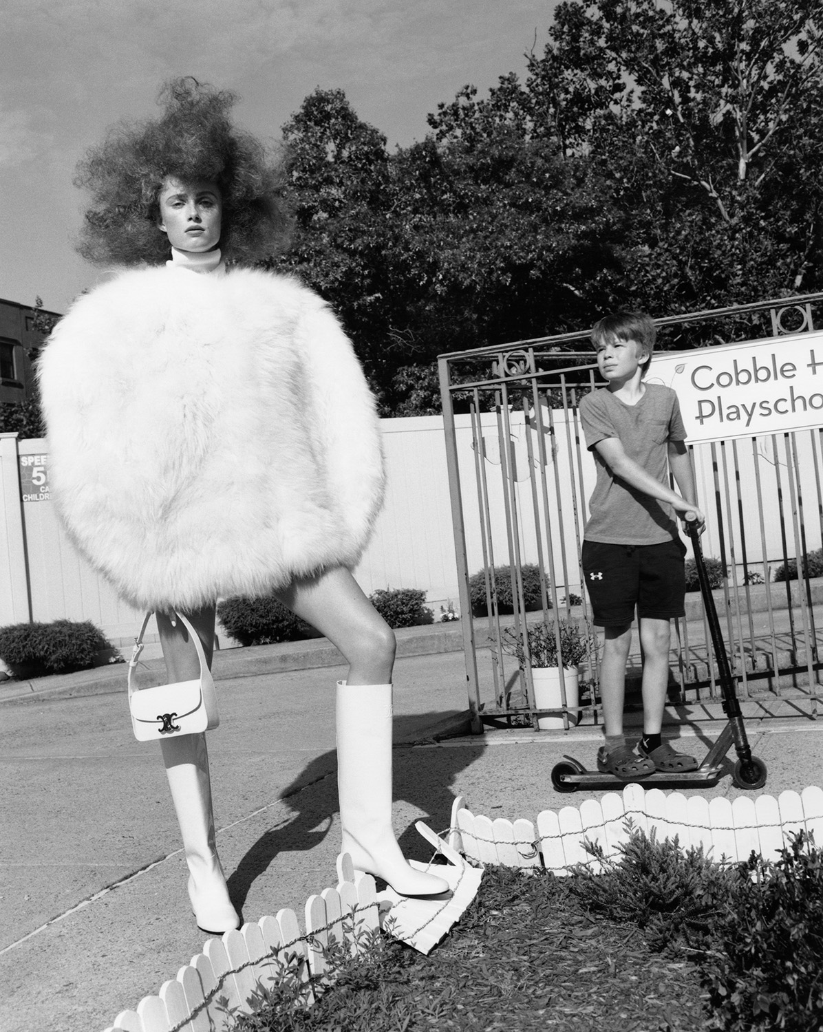 Fashion shot of a model wearing a large white fur coat and boots, holding a purse, alongside a child on a scooter on a sunny day.