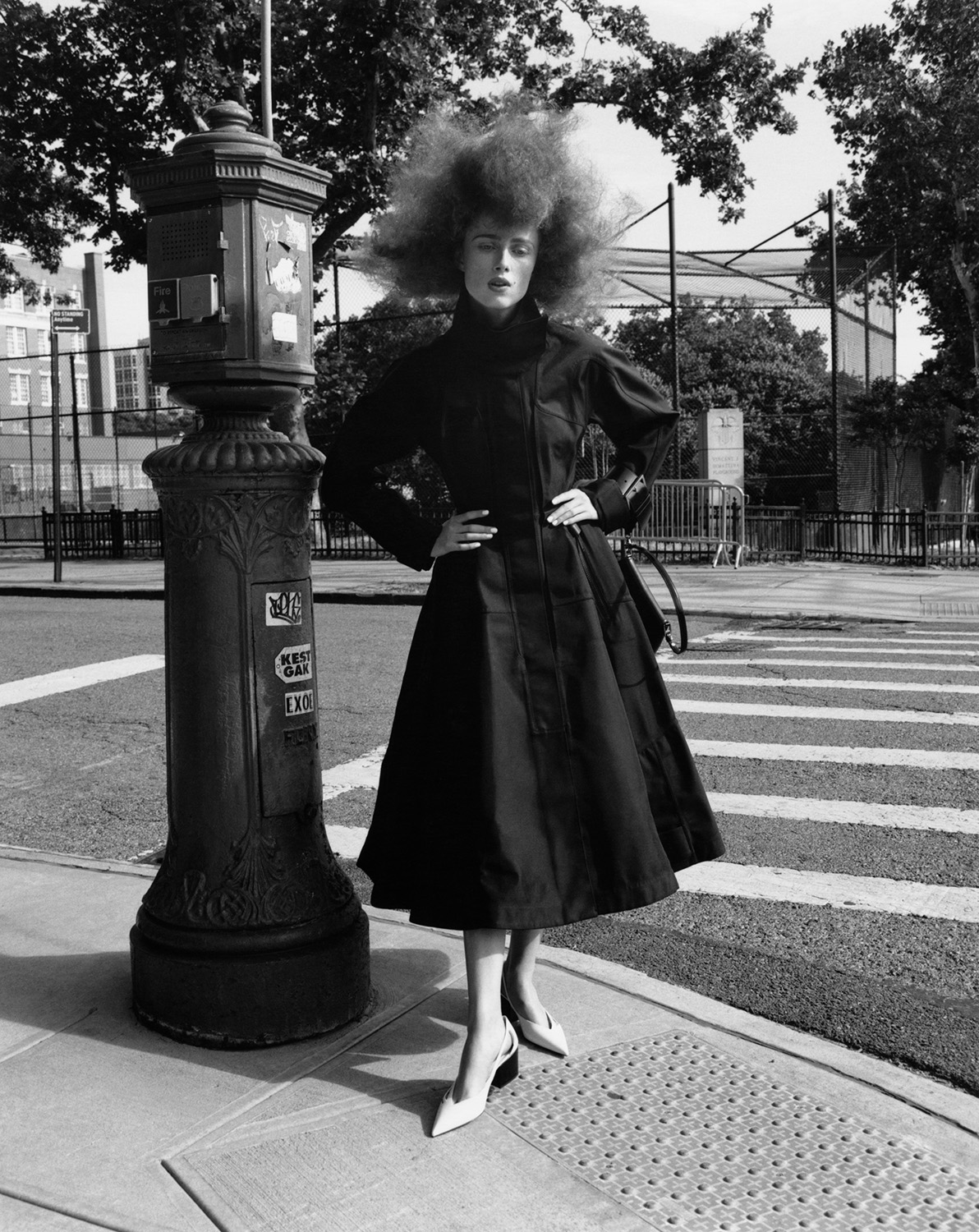 A full shot captures a woman in a dark coat and voluminous hair standing on a city sidewalk next to a vintage-style utility pole.
