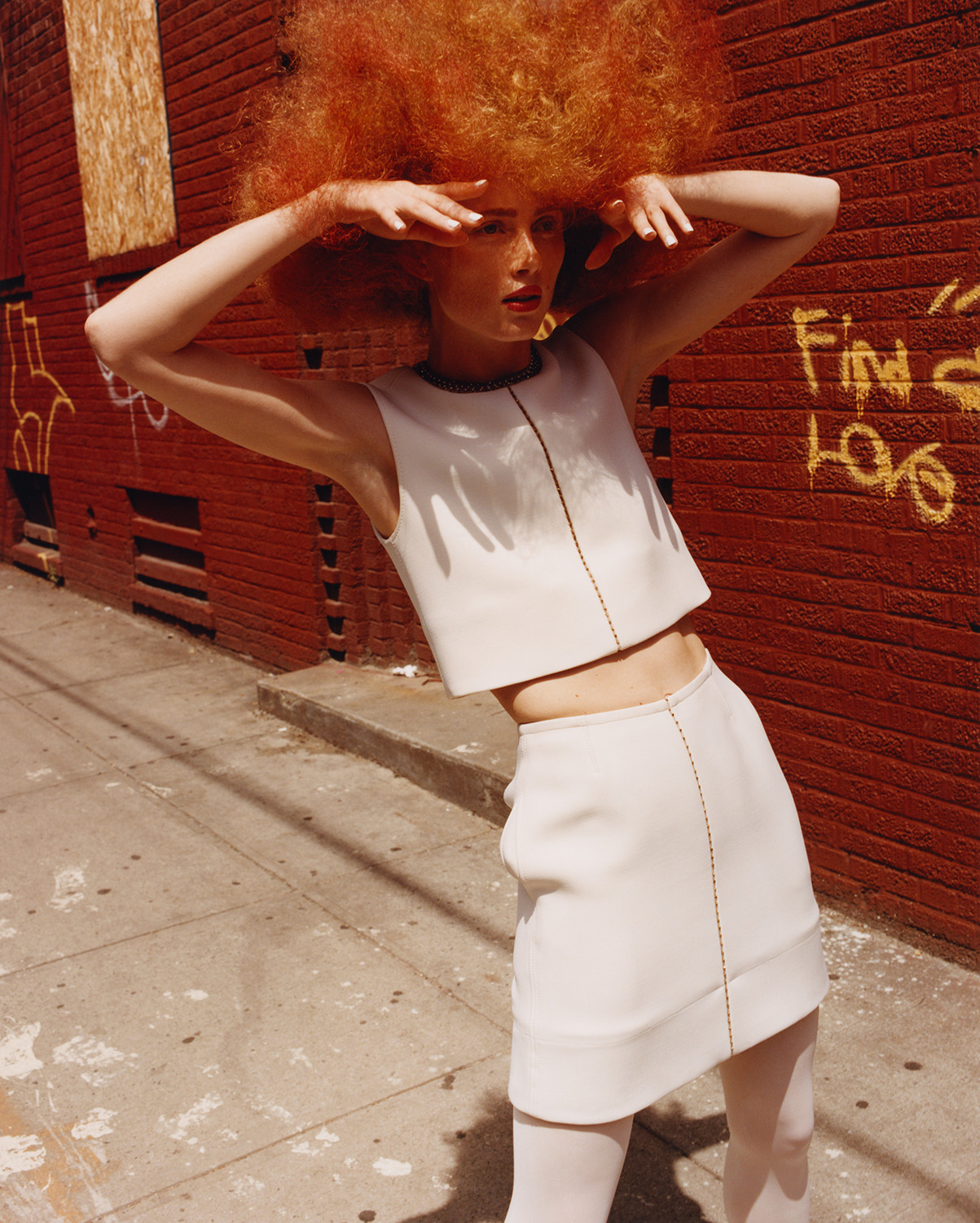 Woman in a white two-piece outfit poses against a red brick wall with graffiti, casting a shadow on a gray sidewalk in an urban setting.