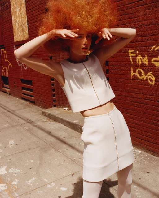 Woman in a white two-piece outfit poses against a red brick wall with graffiti, casting a shadow on a gray sidewalk in an urban setting.
