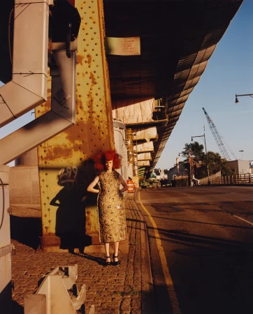 Model with voluminous red hair standing beneath an industrial elevated bridge wearing a metallic embroidered dress