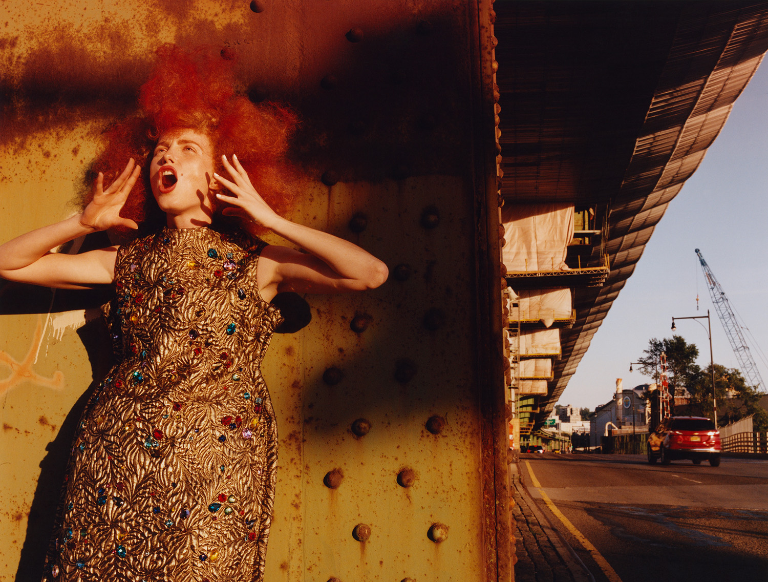 Fashion photo of a woman in a beaded gold dress with red hair, posing expressively under a bridge in an urban outdoor setting.