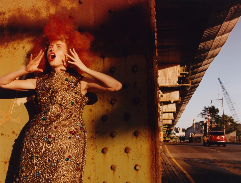 Fashion photo of a woman in a beaded gold dress with red hair, posing expressively under a bridge in an urban outdoor setting.