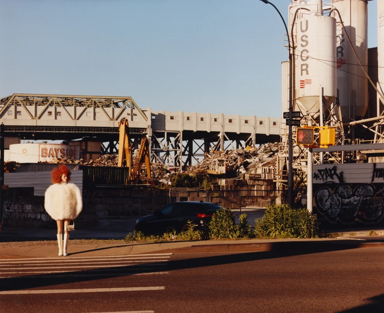 A woman with red hair and a white outfit standing on a zebra crossing, with industrial buildings and a street in the background.