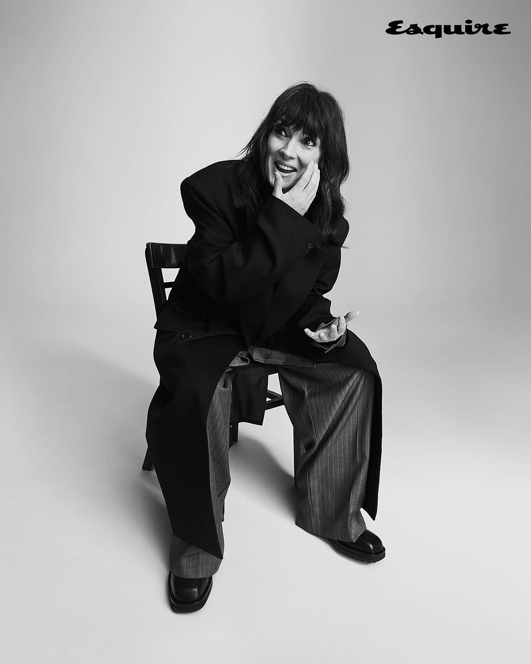Monochrome portrait of a woman with bangs seated on a dark chair wearing an oversized dark coat and patterned trousers.