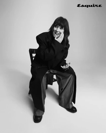 Monochrome portrait of a woman with bangs seated on a dark chair wearing an oversized dark coat and patterned trousers.