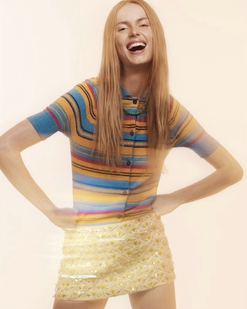 A cheerful young woman in a colorful striped top and beaded yellow skirt laughs with hands on her hips against a light background.