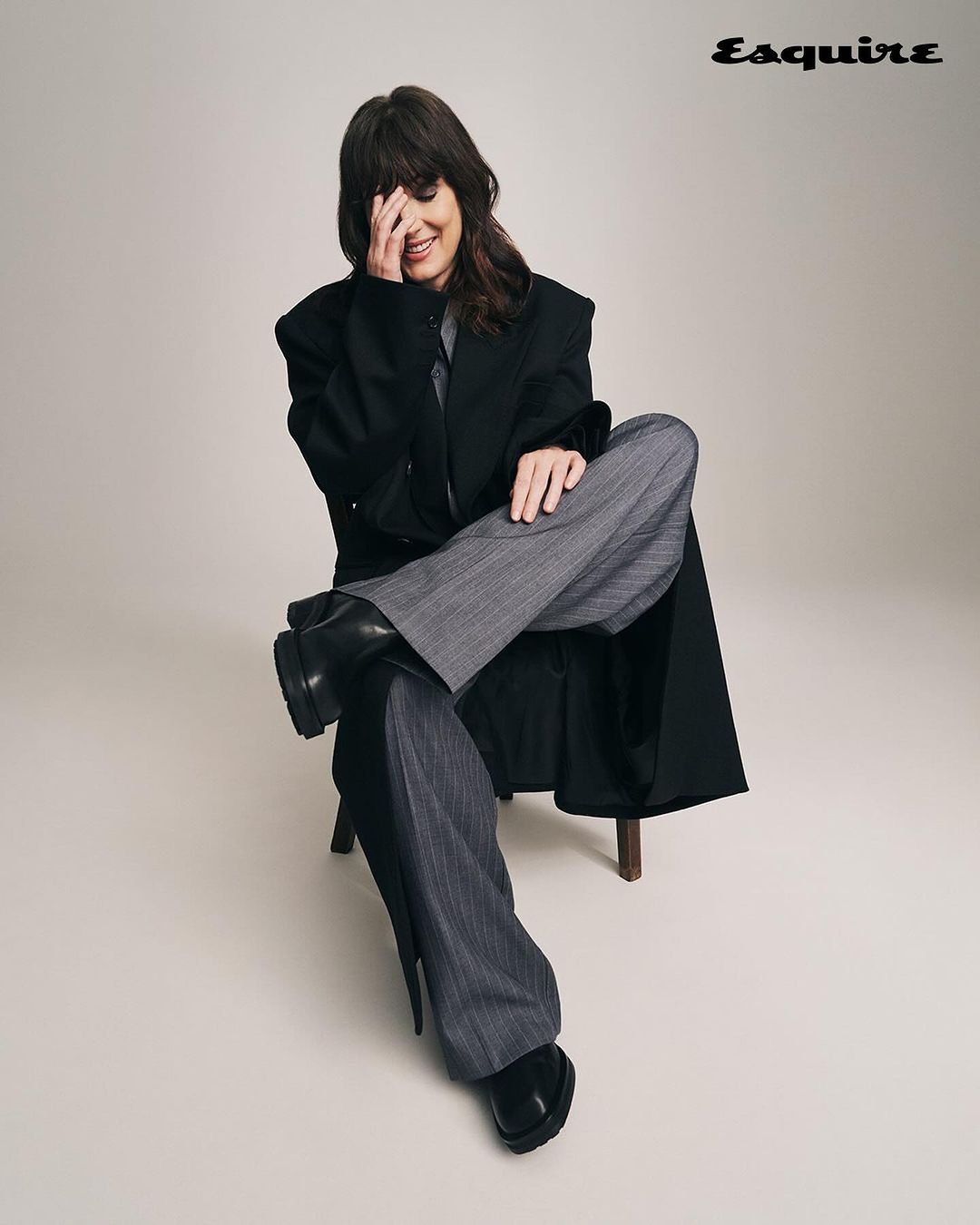 Woman in oversized black coat and grey pinstripe pants sitting on wooden chair in a studio, smiling with hand covering part of her face.