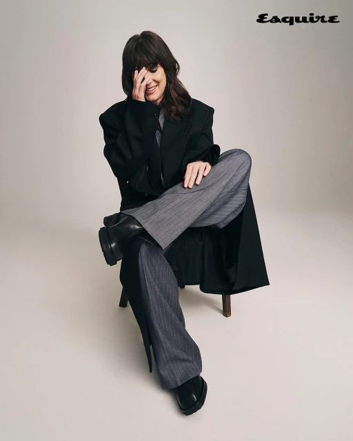 Woman in oversized black coat and grey pinstripe pants sitting on wooden chair in a studio, smiling with hand covering part of her face.