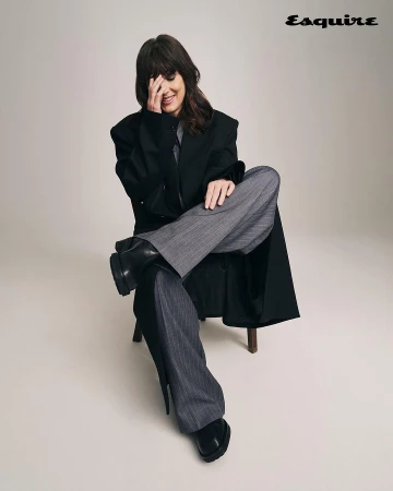 Woman in oversized black coat and grey pinstripe pants sitting on wooden chair in a studio, smiling with hand covering part of her face.