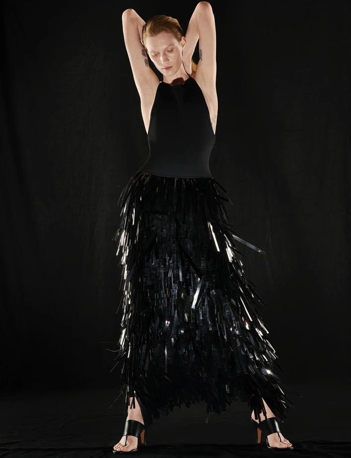 Model in black fringe skirt and halter top posing with arms raised against dark background