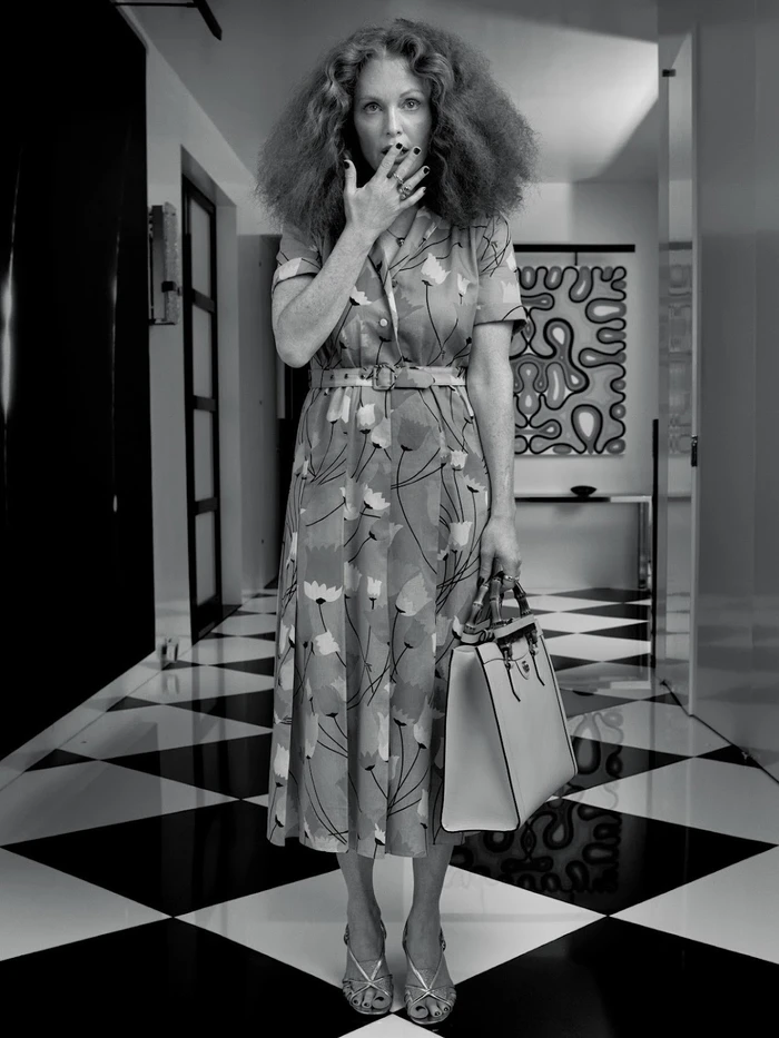 Woman in floral dress with voluminous hair holding a handbag in a hallway with checkered floor by Francelle Daly