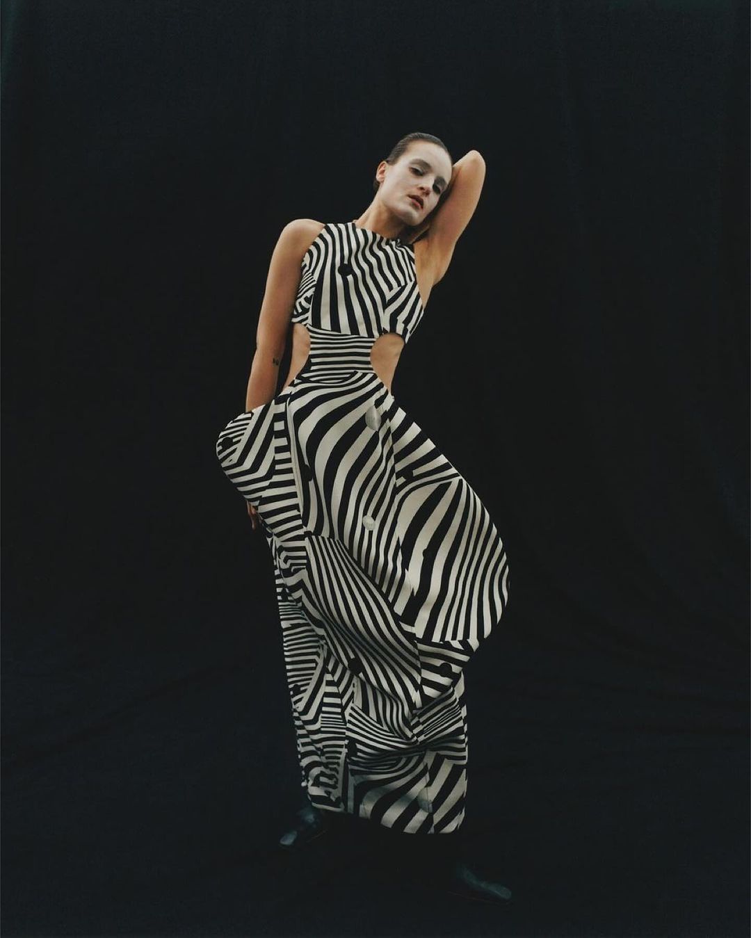 A stylish adult woman in a zebra-striped dress poses in a studio. The woman stands in front of a dark background.