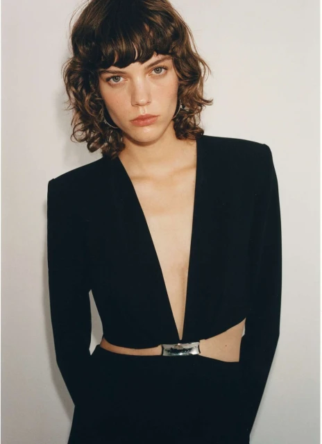 Young woman with brown wavy hair, wearing a deep v-neck black dress with silver belt, posing indoors in front of a plain white wall.