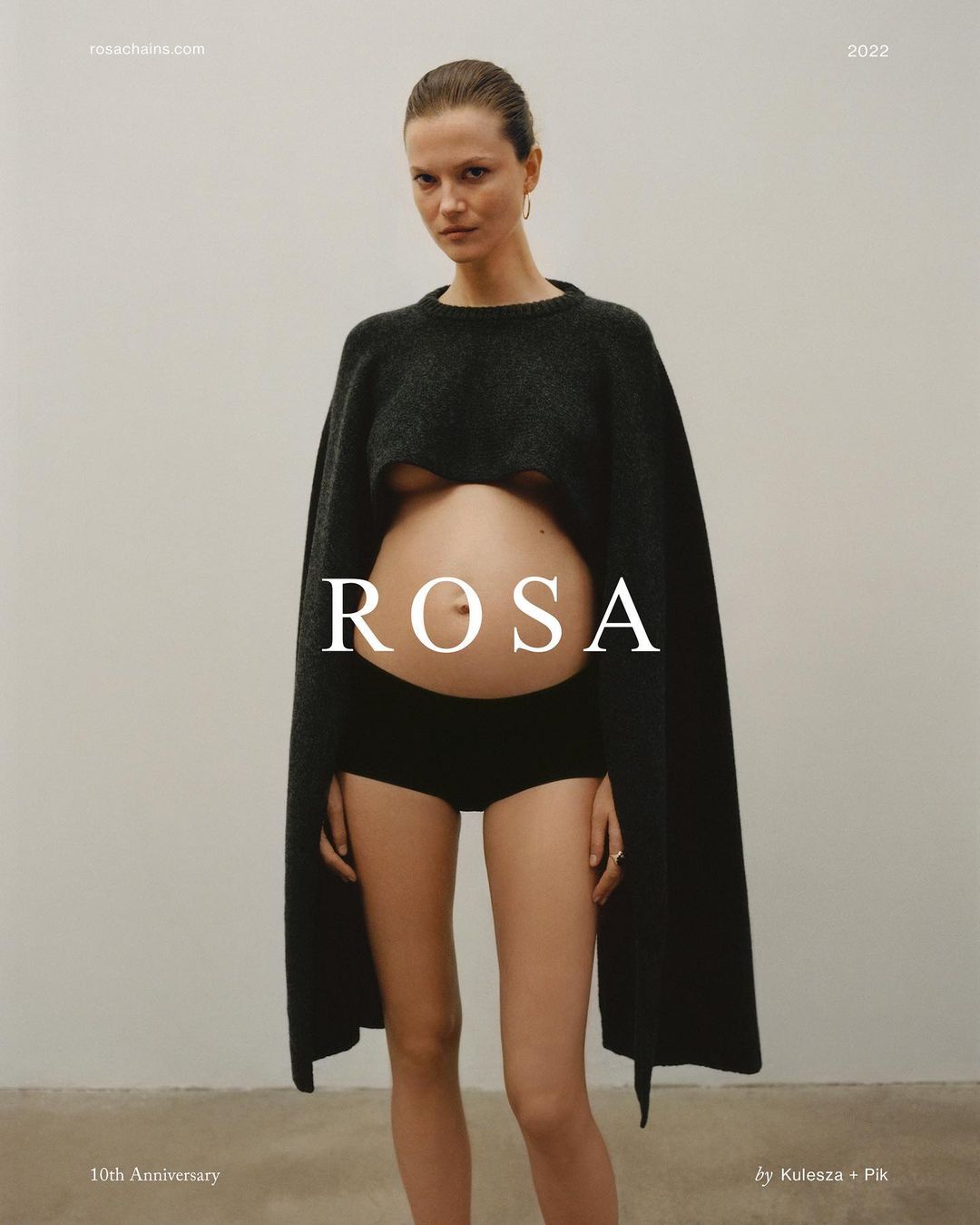 Portrait of pregnant woman in black knitwear, black bottoms, and minimal makeup against a neutral background, showcasing fashion.