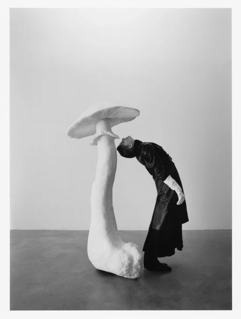 Black and white portrait of a fashion model in a black coat leaning over an oversized mushroom in a minimalist studio setting.