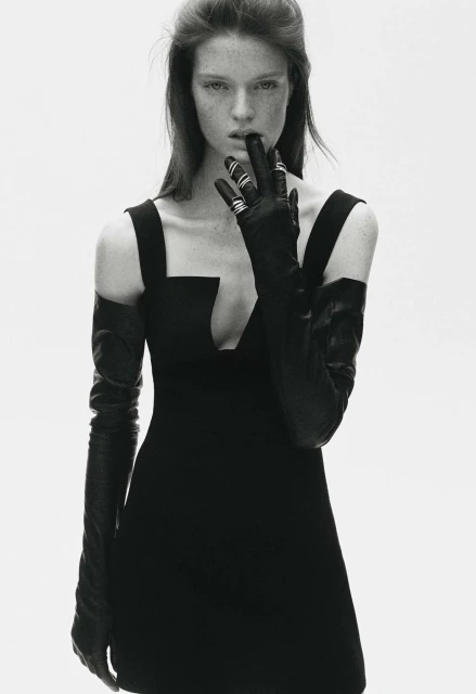 Monochromatic fashion shot: A woman with fair skin poses in a black dress and gloves against a bright white backdrop.