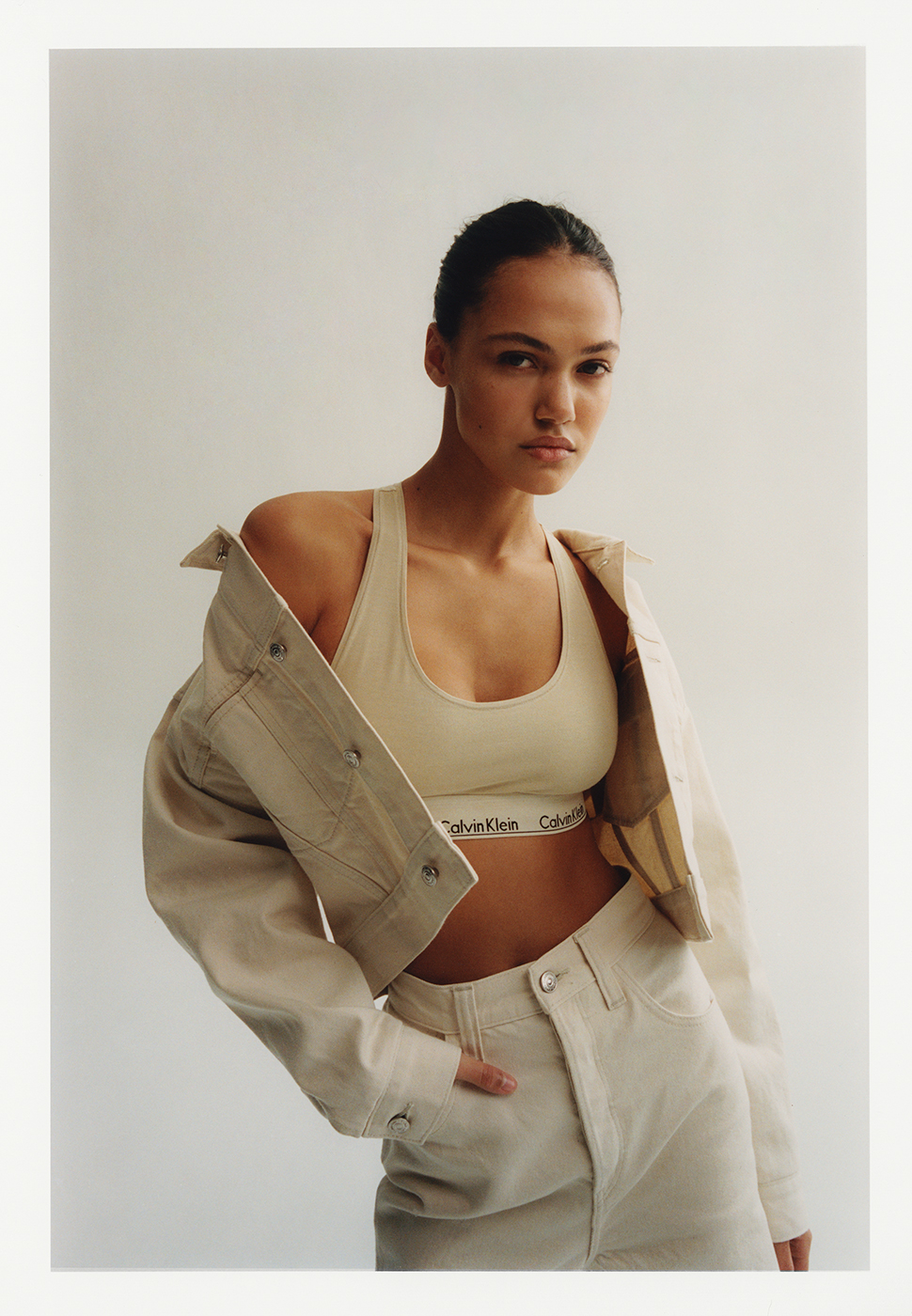 Model in cream denim jacket and matching bralette posing against a solid white background in a portrait photograph