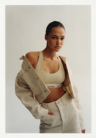 Model in cream denim jacket and matching bralette posing against a solid white background in a portrait photograph