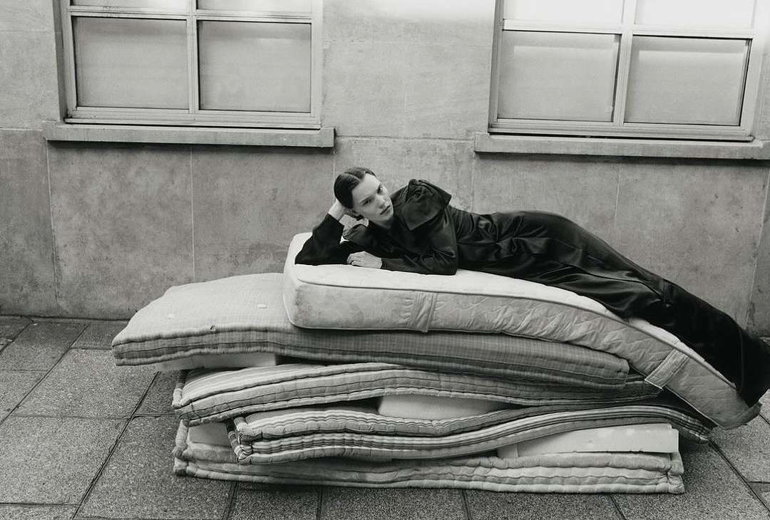 Monochrome fashion portrait of a woman reclining on a stack of mattresses, set against a stark concrete wall and window.