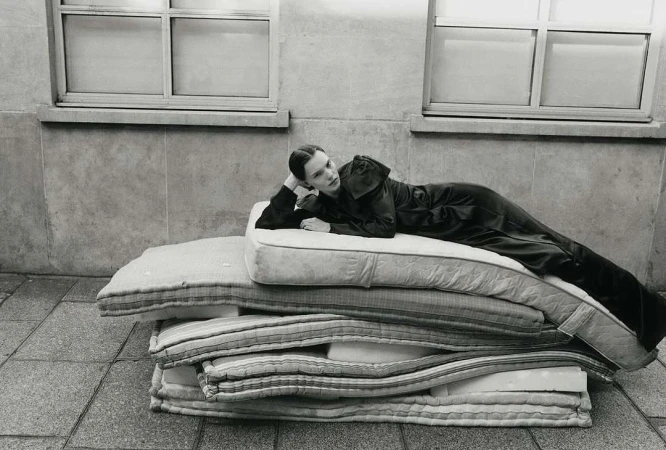 Monochrome fashion portrait of a woman reclining on a stack of mattresses, set against a stark concrete wall and window.