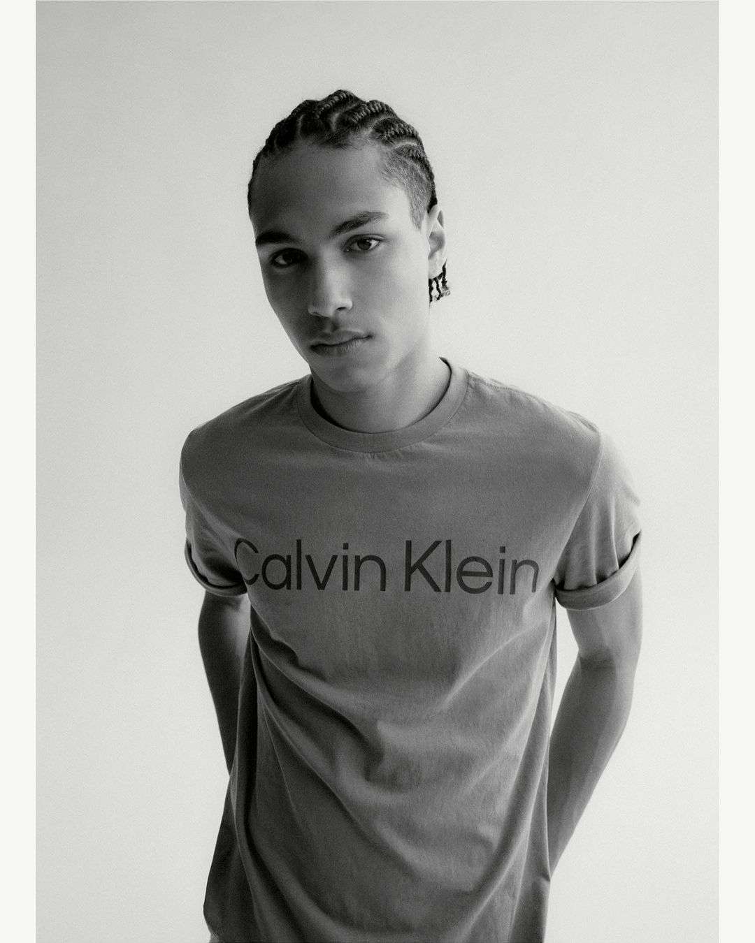 Monochrome portrait of a young adult male with braided hair, wearing a t-shirt with a visible 'Calvin Klein' logo against a white backdrop.