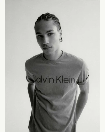 Monochrome portrait of a young adult male with braided hair, wearing a t-shirt with a visible 'Calvin Klein' logo against a white backdrop.