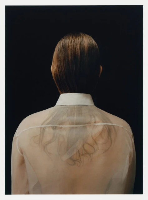 Back view of an adult with short brown hair, wearing a sheer shirt with a white collar, set against a solid black background.
