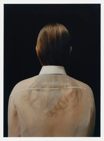 Back view of an adult with short brown hair, wearing a sheer shirt with a white collar, set against a solid black background.