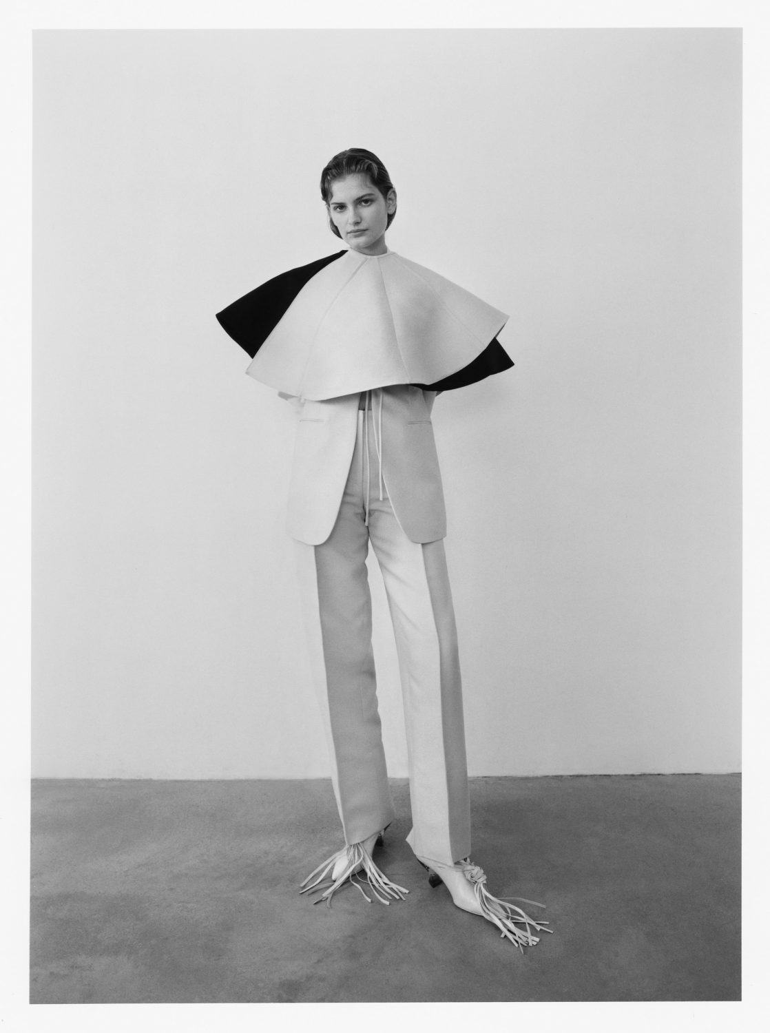 Fashion model posing in a studio with a cream and black layered cape, jacket, and two-tone pants with fringed heels against a grey backdrop.