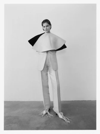 Fashion model posing in a studio with a cream and black layered cape, jacket, and two-tone pants with fringed heels against a grey backdrop.
