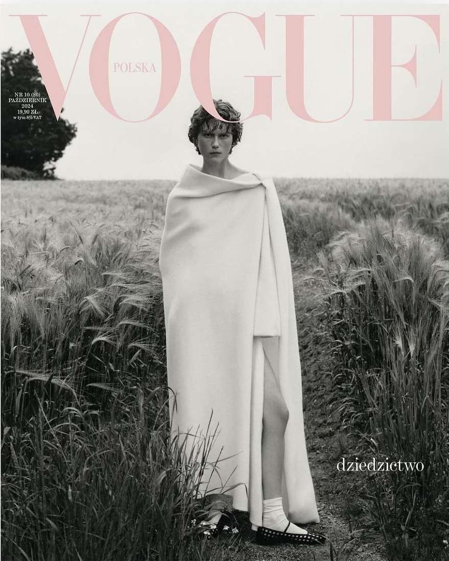 Model wrapped in a white blanket standing in a tall grass field under an overcast sky for a magazine cover.