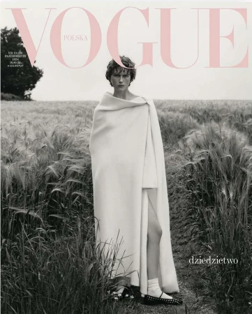 Model wrapped in a white blanket standing in a tall grass field under an overcast sky for a magazine cover.