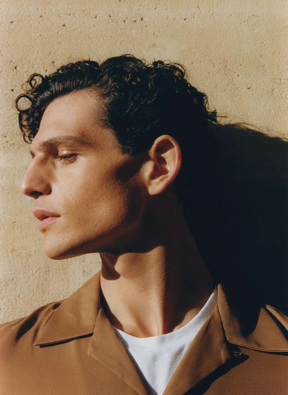 Portrait of an adult male with curly hair, wearing a brown collared shirt and a white t-shirt, with a neutral facial expression.