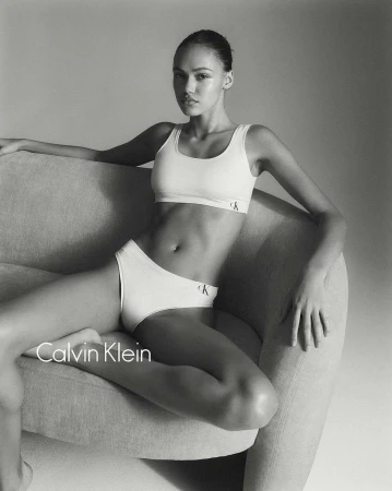 Woman in white Calvin Klein underwear and bra top poses on a grey couch in a studio. Fashion portrait with neutral grey background.