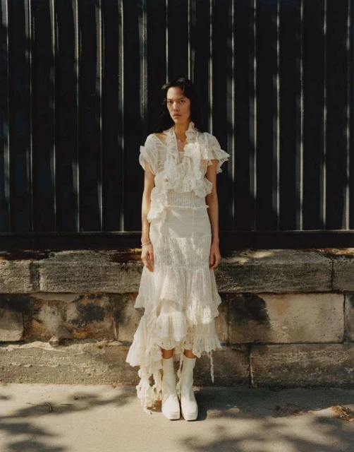 Fashionable woman poses in a cream lace dress and platform boots against a textured wall, blending modern style with classic elegance.