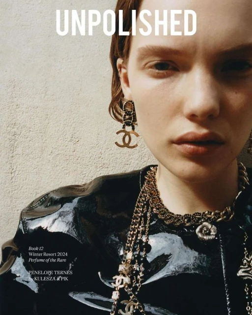 Close-up of a red-haired woman wearing Chanel earrings, a black patent leather jacket, and multiple gold necklaces in a studio setting.