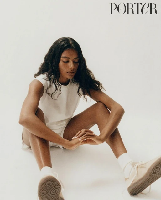 A young woman poses in a white top and shorts, seated indoors with sneakers, against a bright backdrop for a fashion editorial.