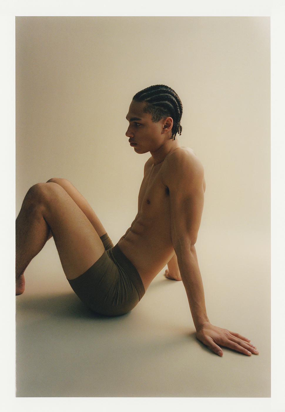 An adult male model is shown in a studio setting wearing boxer briefs. The man is sitting on the floor and looking to the side.