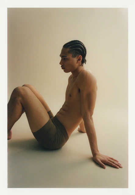 An adult male model is shown in a studio setting wearing boxer briefs. The man is sitting on the floor and looking to the side.