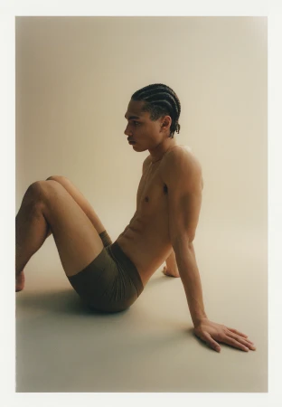 An adult male model is shown in a studio setting wearing boxer briefs. The man is sitting on the floor and looking to the side.