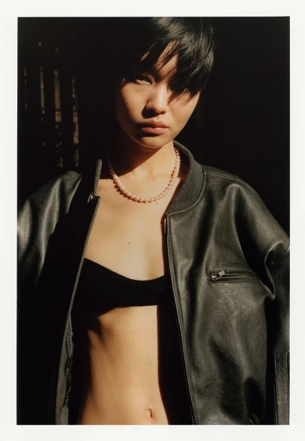Close-up portrait of an Asian woman in a black leather jacket and black bra top, set against a dark background and natural light.