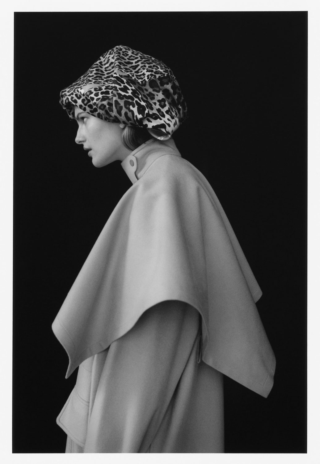 Black-and-white portrait of a woman in profile wearing a leopard-print hat and cream-colored cape or coat against a black backdrop.