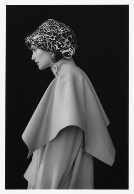 Black-and-white portrait of a woman in profile wearing a leopard-print hat and cream-colored cape or coat against a black backdrop.
